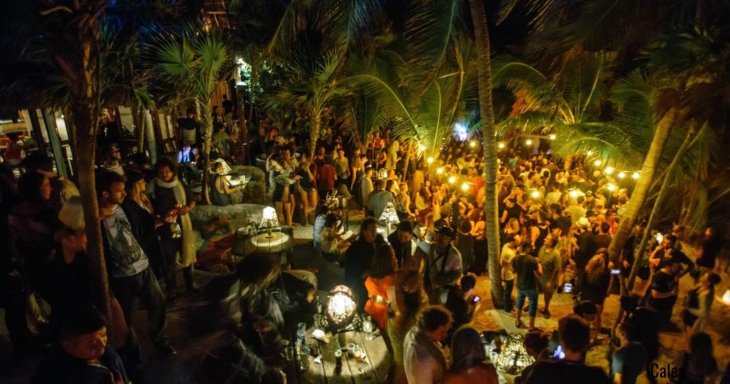 Una vista aérea de una fiesta nocturna al aire libre con una gran multitud de personas. El área está iluminada por numerosas luces cálidas y guirnaldas, creando un ambiente festivo. Hay muchas palmeras que se elevan sobre la multitud, con sus hojas apenas visibles contra el cielo nocturno. Algunas personas están de pie, otras sentadas en mesas redondas, y algunos tienen bebidas en la mano. La escena sugiere un evento social animado en un clima cálido.