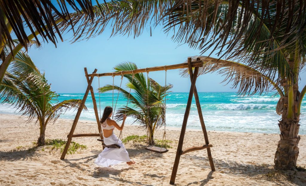 A woman swinging on a wooden swing, facing the turquoise waters and white sands of a serene beach in Tulum, Mexico.