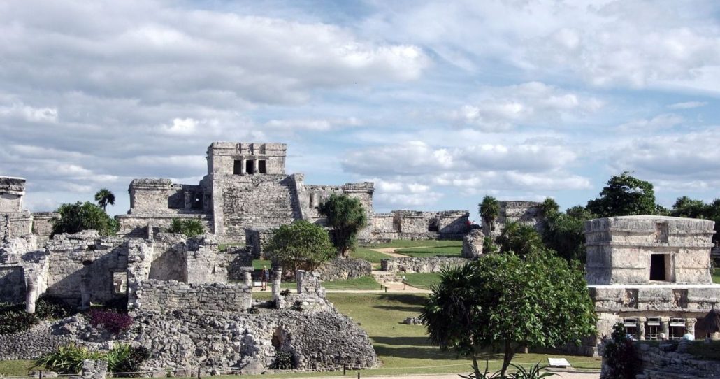 Una vista panorámica de las ruinas mayas de Tulum bajo un cielo parcialmente nublado. Se ven varias estructuras de piedra antiguas, algunas con aberturas rectangulares y otras en ruinas. La vegetación verde, incluyendo árboles y arbustos, está dispersa entre las estructuras de piedra, y hay áreas de césped bien cuidado.
