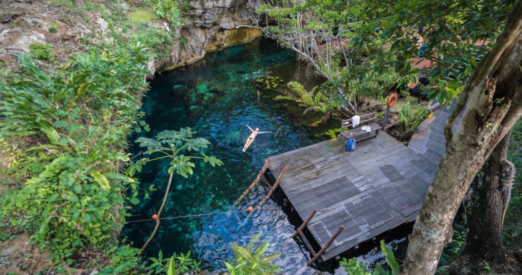 Una vista aérea de un cenote con aguas claras de color turquesa. Una persona está flotando de espaldas en el agua. A la derecha, un muelle de madera se extiende hacia el agua con una mesa de picnic y pertenencias sobre él. El cenote está rodeado de densa vegetación verde y formaciones rocosas.
