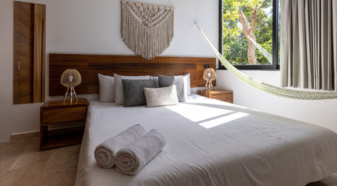 Bright room at Hotel Calea Tulum with natural wood bed, sand-toned decor, and a window view of lush trees — perfect stay after getting to Tulum.