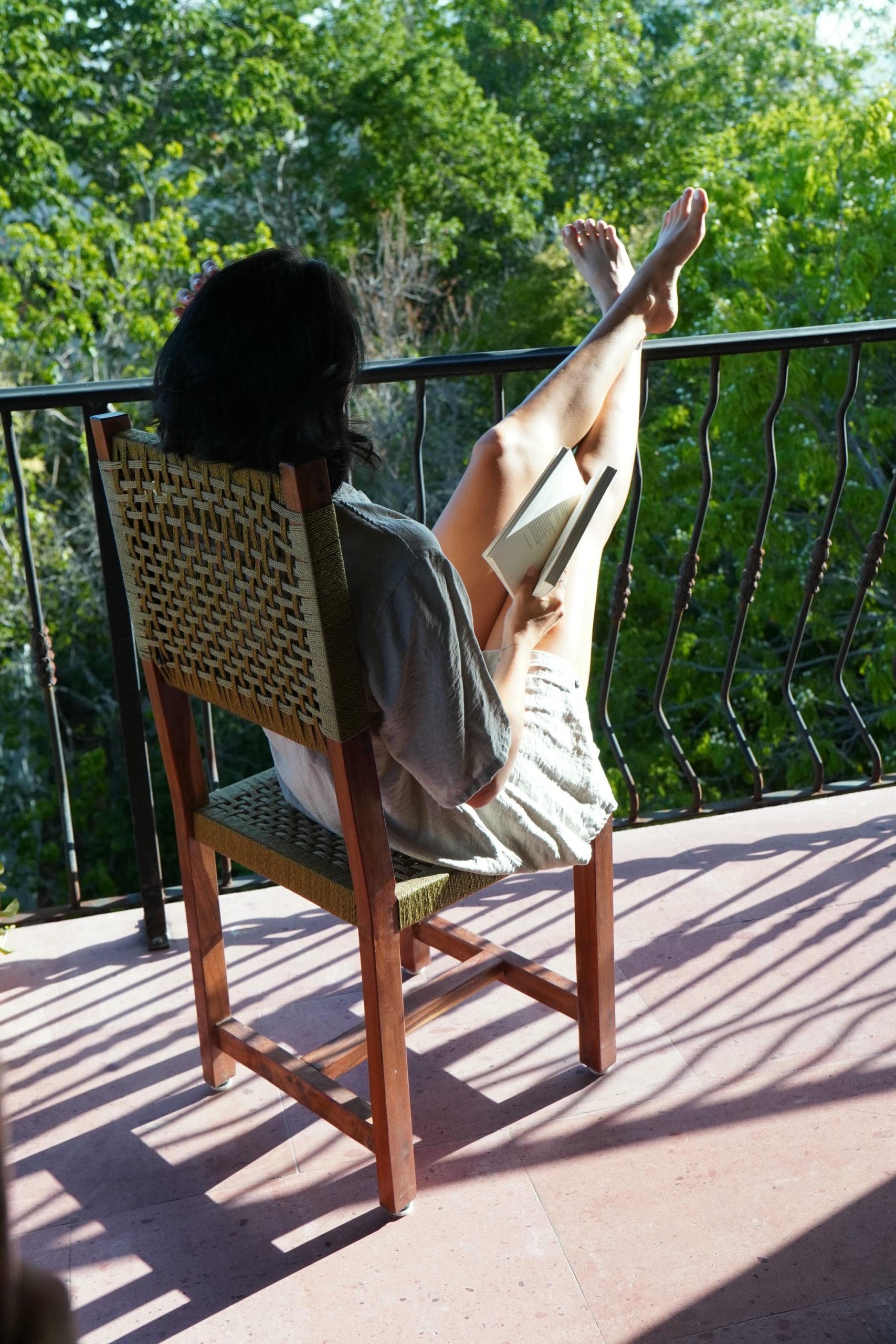Una mujer vista desde atrás, sentada en una silla de madera en un balcón soleado de Calea Tulum, con las piernas apoyadas en la barandilla mientras lee un libro. Al fondo se ve un paisaje frondoso y verde.