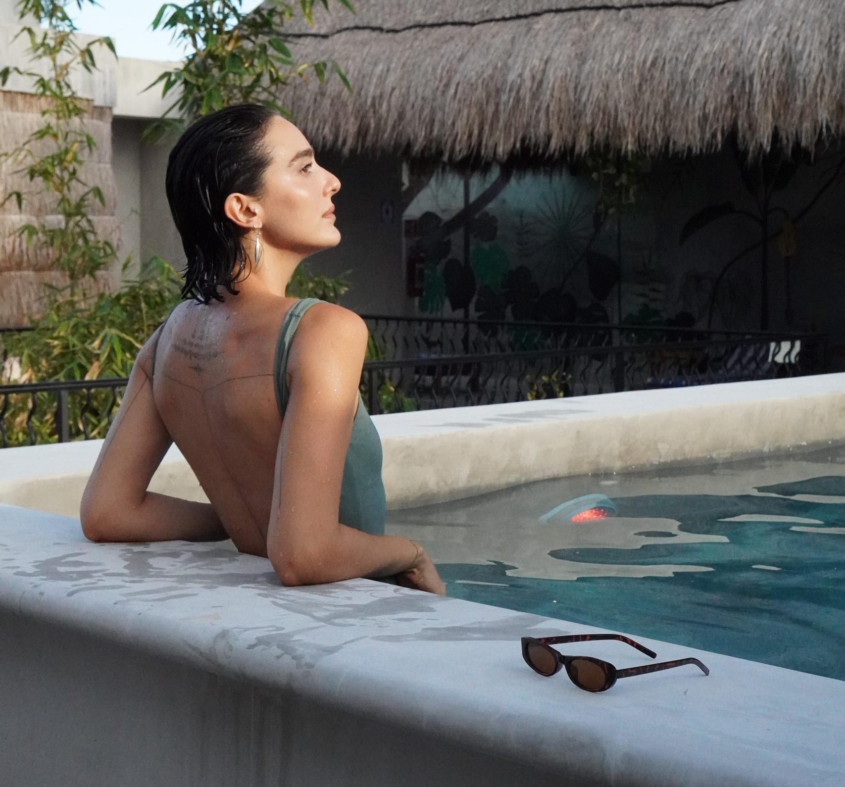 Una mujer con cabello oscuro y húmedo, luciendo un traje de baño color verde oliva, está recostada en el borde de la piscina de Calea Tulum. Tiene un tatuaje en la espalda. En primer plano, unas gafas de sol de carey descansan en el borde de la piscina. El fondo muestra un techo de paja y vegetación.